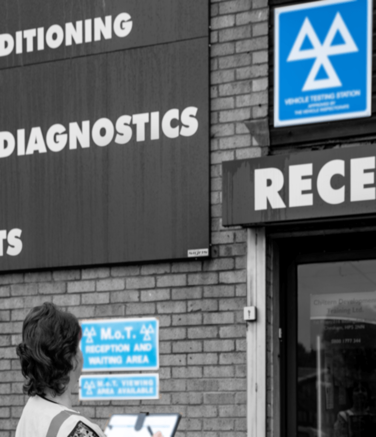 WOman in hi-vis jacket with clipboard looking at MOT Testing Station signage